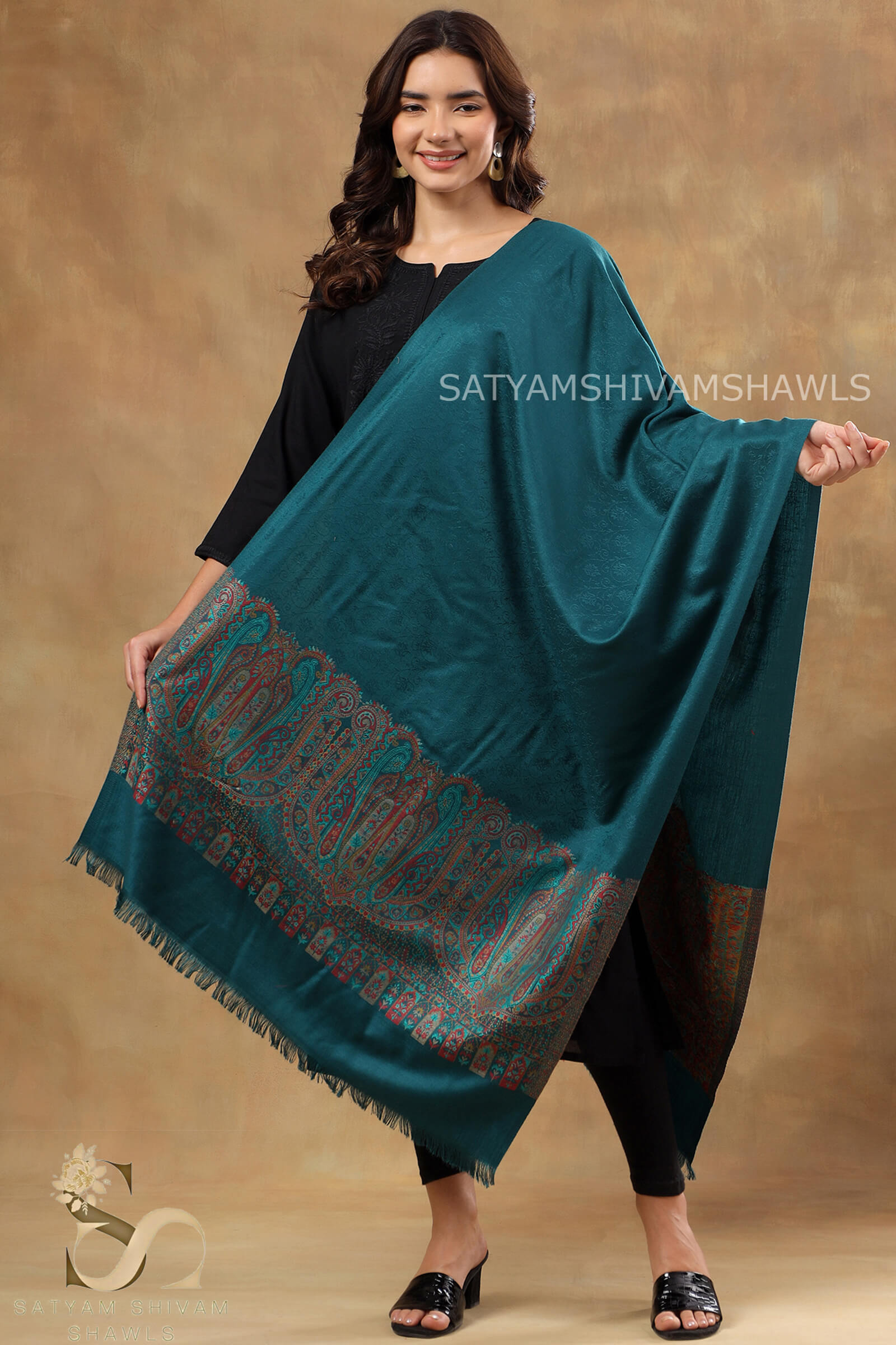 Woman holding a teal shawl with intricate patterns against a brown background, branded 'Satyam Shivam Shawls'.